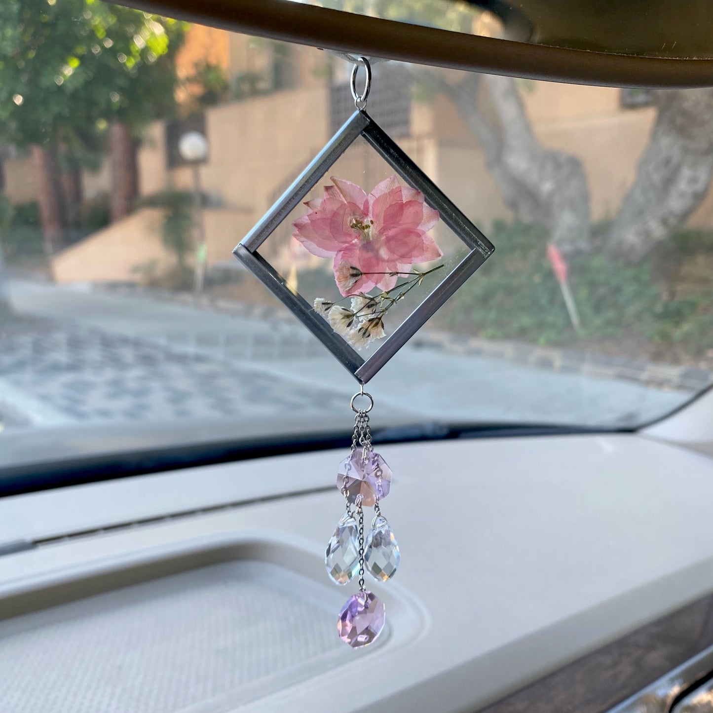 A handmade sun catcher with a floral design, featuring a pink flower and clear crystals, hanging from a car's rearview mirror.